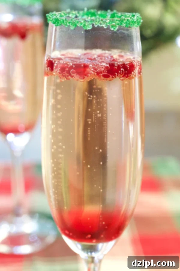 Close-up of a vibrant Christmas Champagne Cocktail featuring layers of ruby-red pomegranate simple syrup, effervescent champagne, and fresh pomegranate seeds. The flute is adorned with a sparkling green sugar rim, capturing the essence of holiday festivity.
