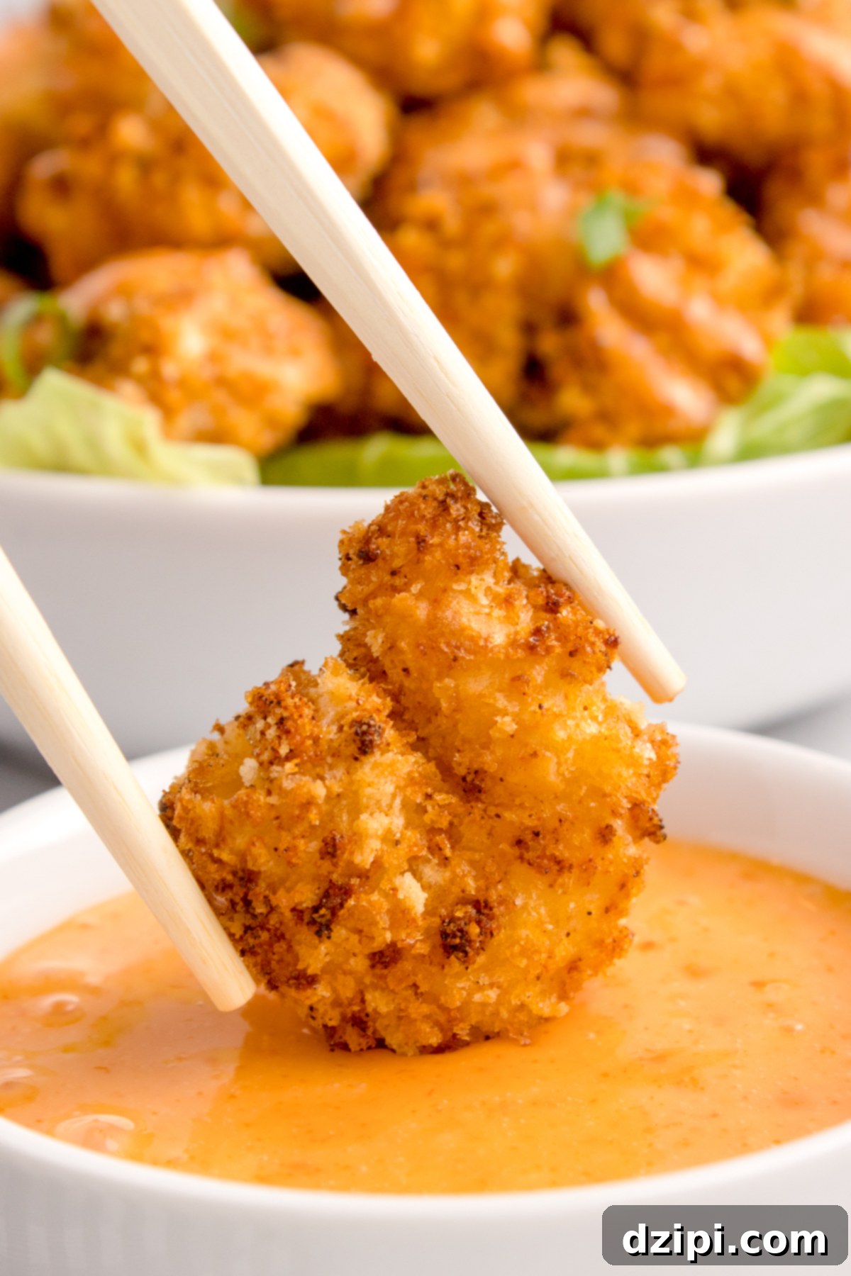 A pair of chopsticks holds a crispy breaded shrimp that's being dipped in a bowl or orange colored bang bang sauce.