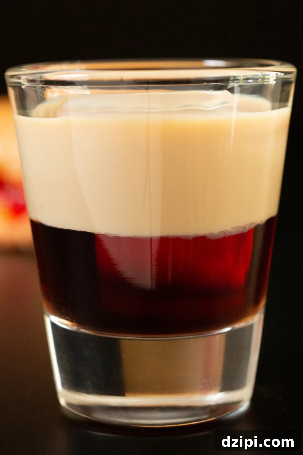 Jam Doughnut Poppers 2 A shot glass is filled with two distinct layers - a vibrant crimson red raspberry liqueur on the bottom, and a creamy beige Irish cream liqueur on top. The perfect Jam Donut Shot for dessert lovers, highlighting its visual appeal.