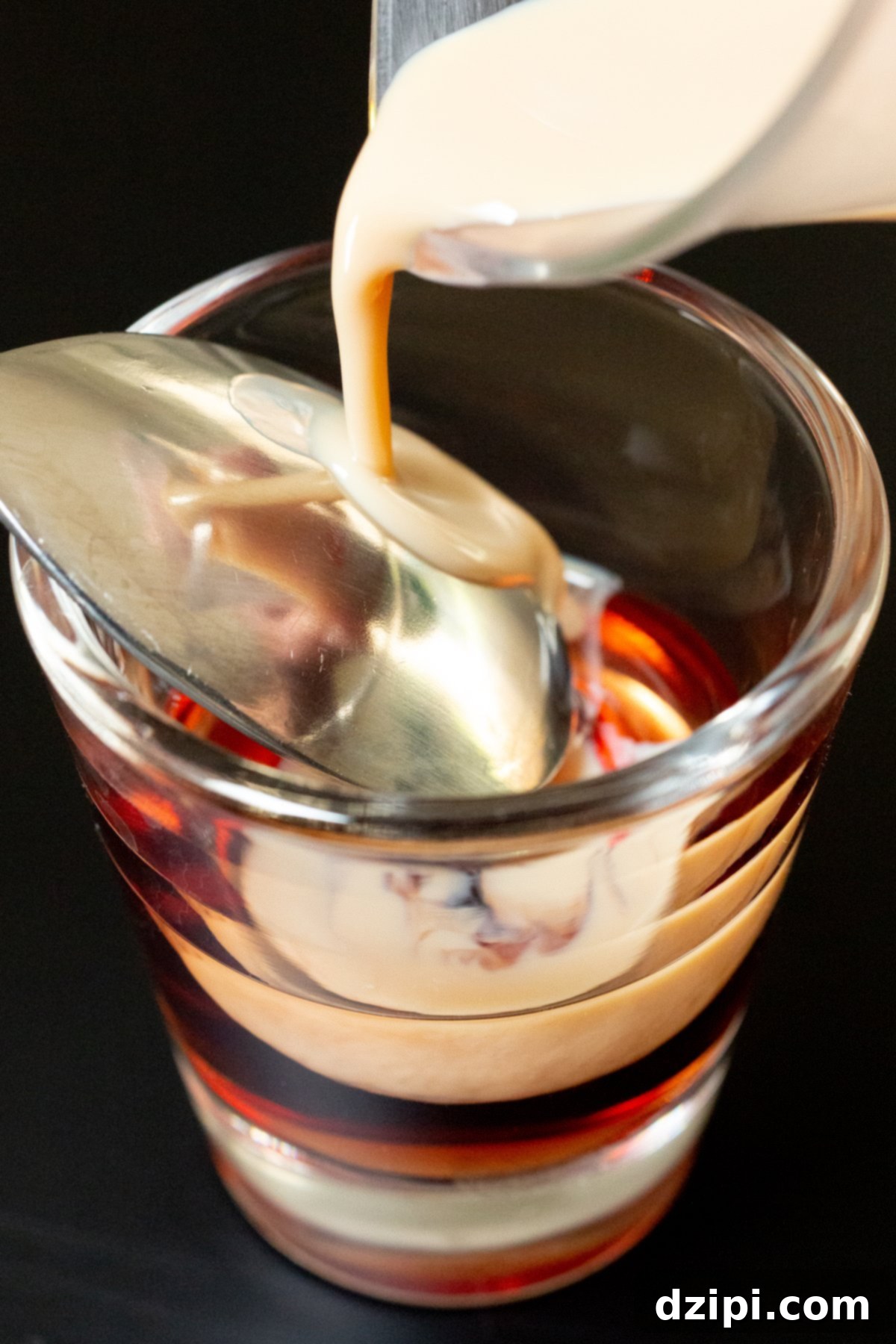 Jam Doughnut Poppers 4 Irish cream liqueur being slowly and carefully poured over the back of a metal bar spoon, which is precisely positioned just above a vibrant red layer of Chambord in a shot glass. This technique ensures distinct and beautiful layering for the Jam Donut Shot.