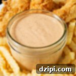 A white dinner plate has a small jar of light pink sauce with visible specs of black peppers and other spices. It is surrounded by crispy chicken tenders and wavy fries.