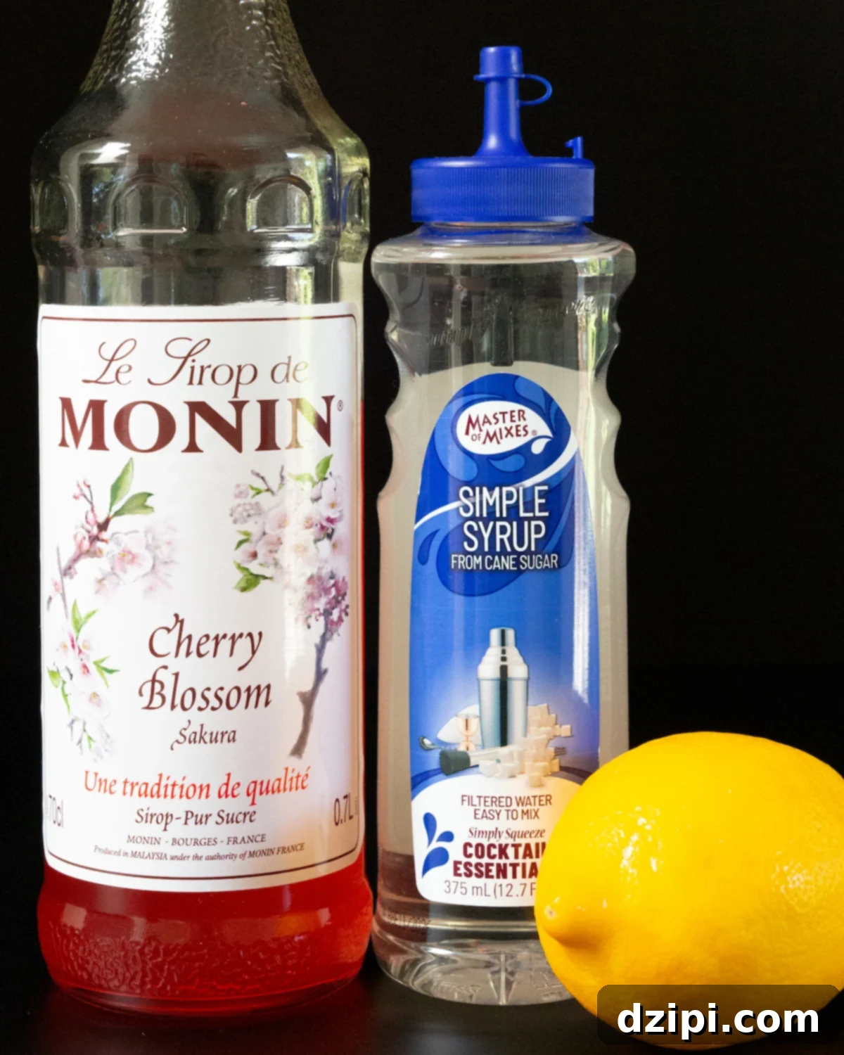 A bottle of cherry blossom syrup, simple syrup, and a lemon on a black background.