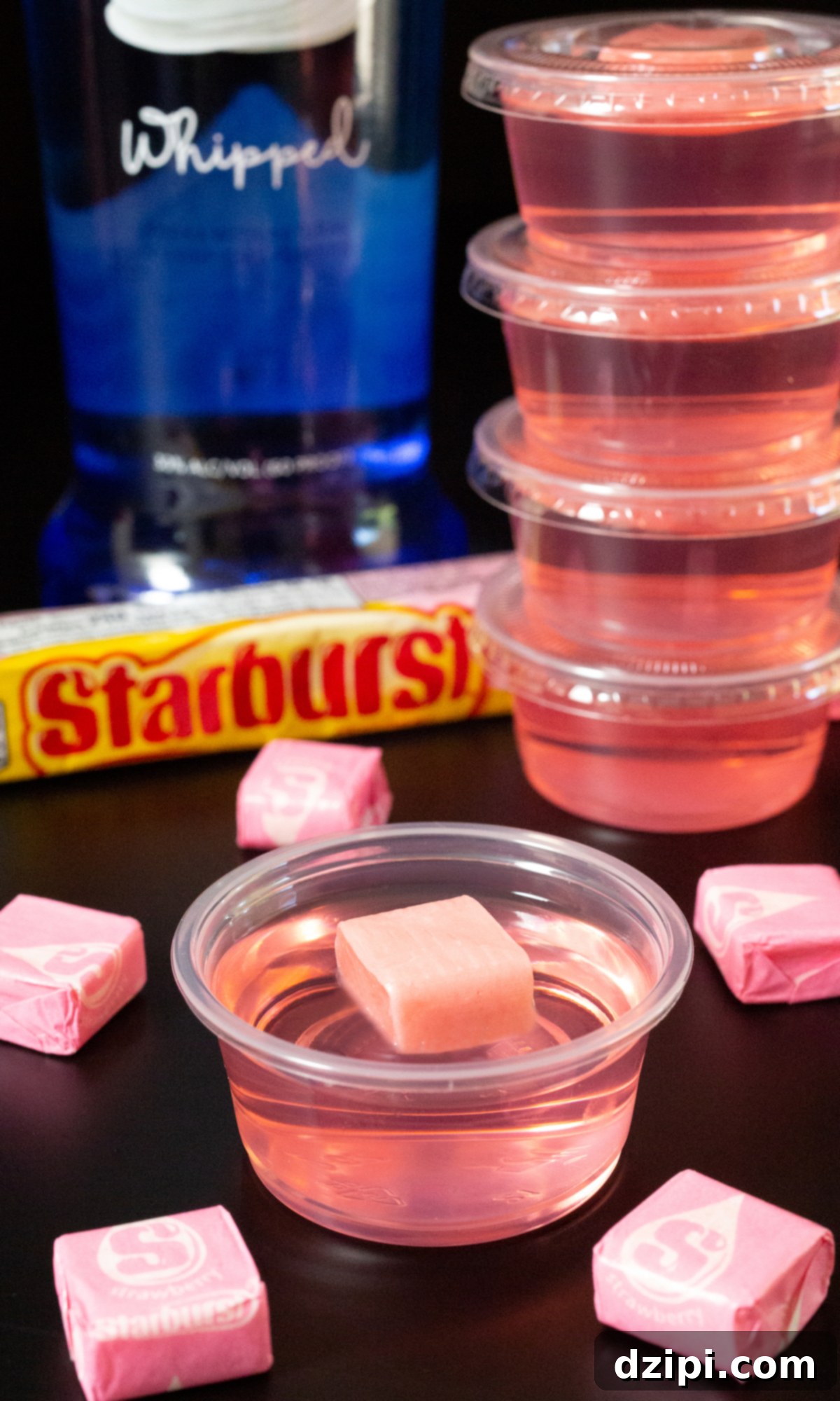 A single pink jello shot topped with an unwrapped pink Starburst, in focus. In the blurred background, more jello shots and a bottle of whipped vodka are visible.