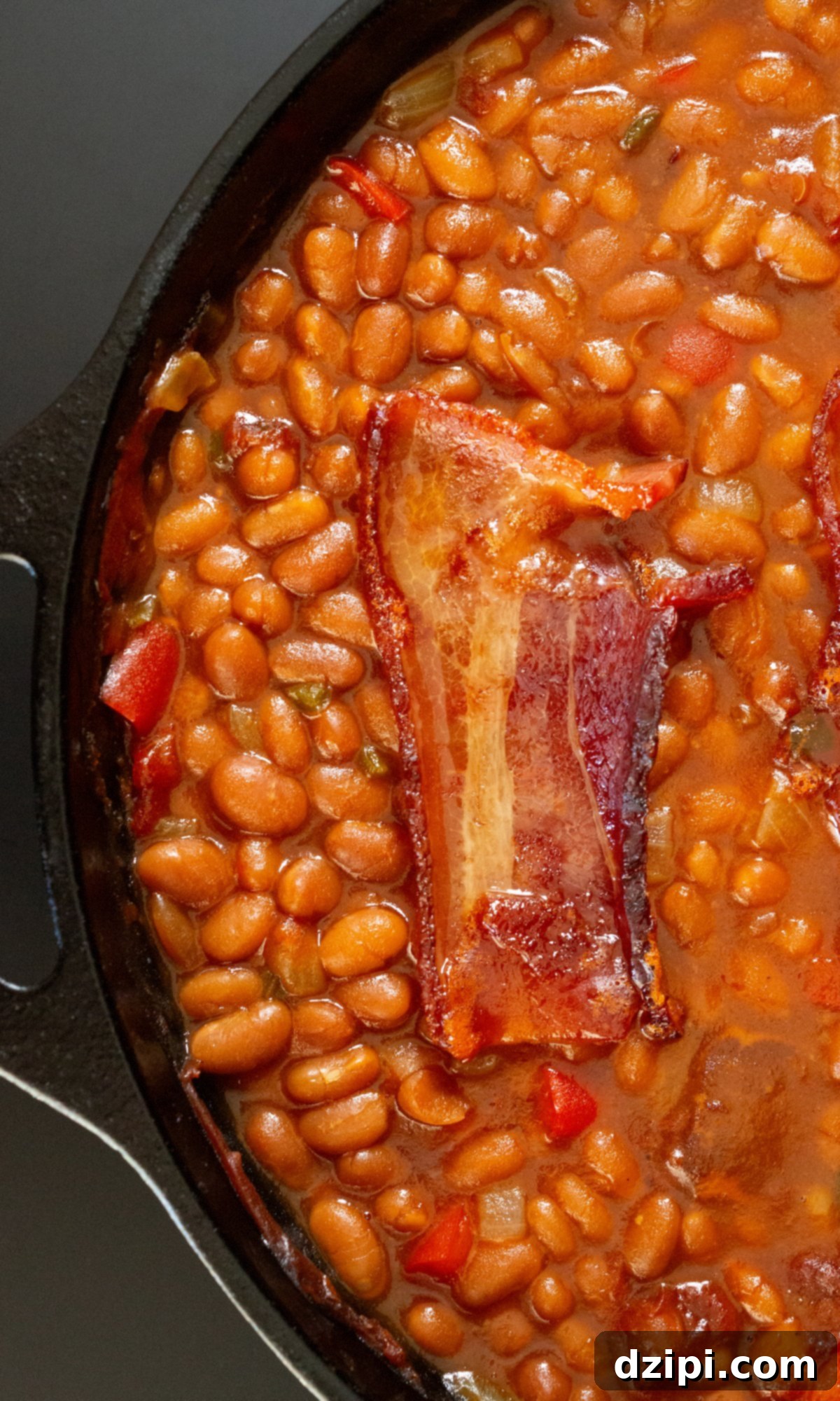 A cast iron skillet is filled with smoked baked beans that have visible chunks of vegetables and onions. Topped with a crispy slice of bacon.