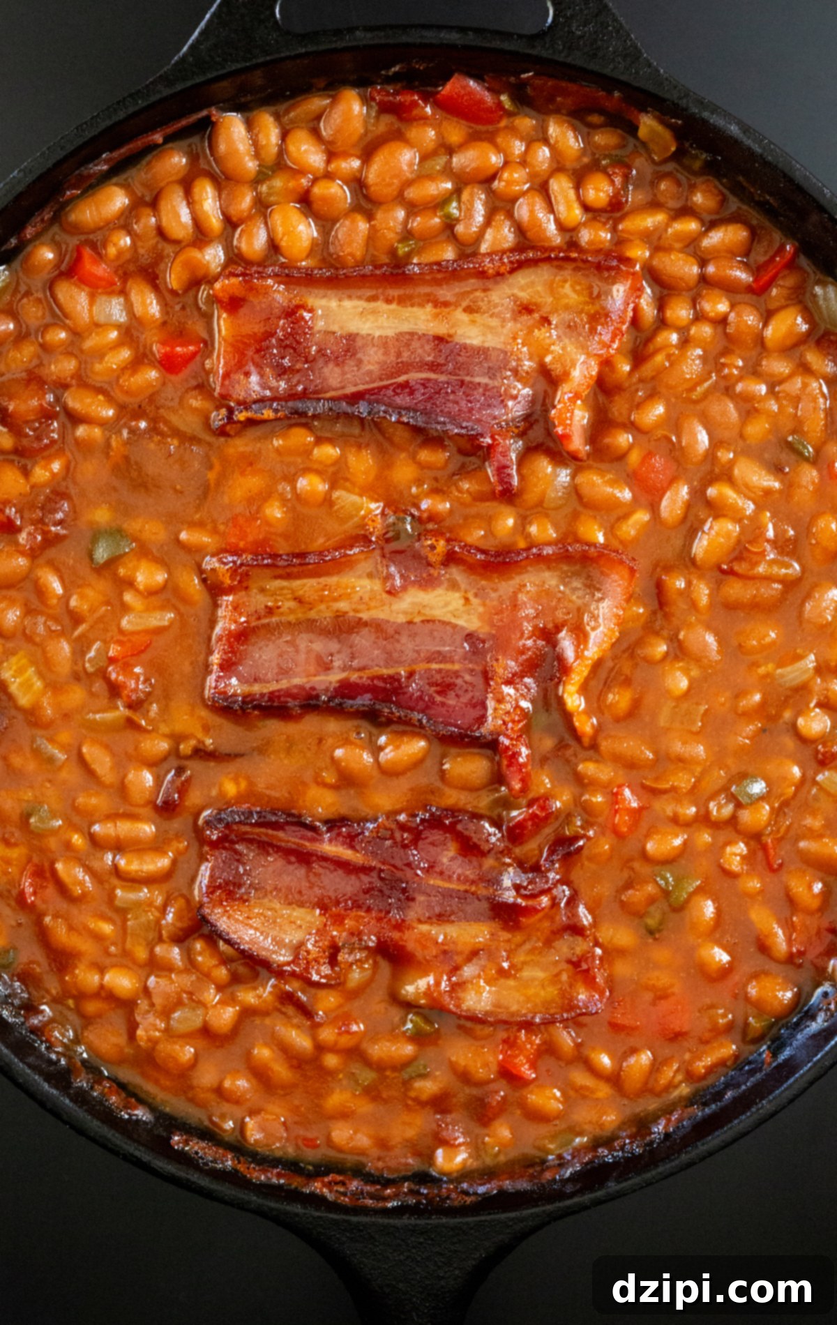 A cast-iron skillet filled with smoked baked beans that are topped with three slices of cooked bacon.