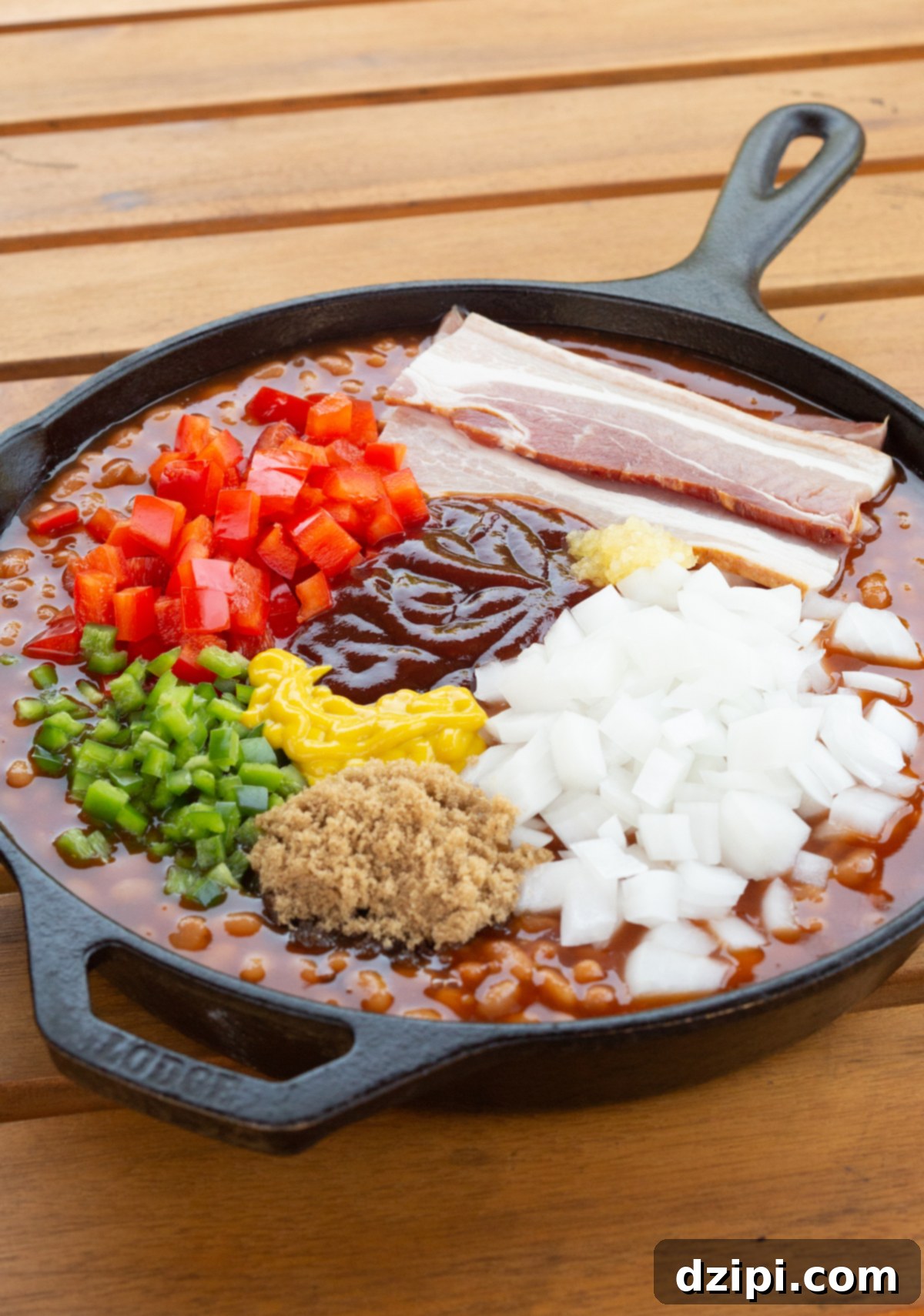 A cast iron skillet that's been filled with baked beans then topped with raw bacon slices, diced red and green pepper, onion, garlic, bbq sauce, mustard, and brown sugar.