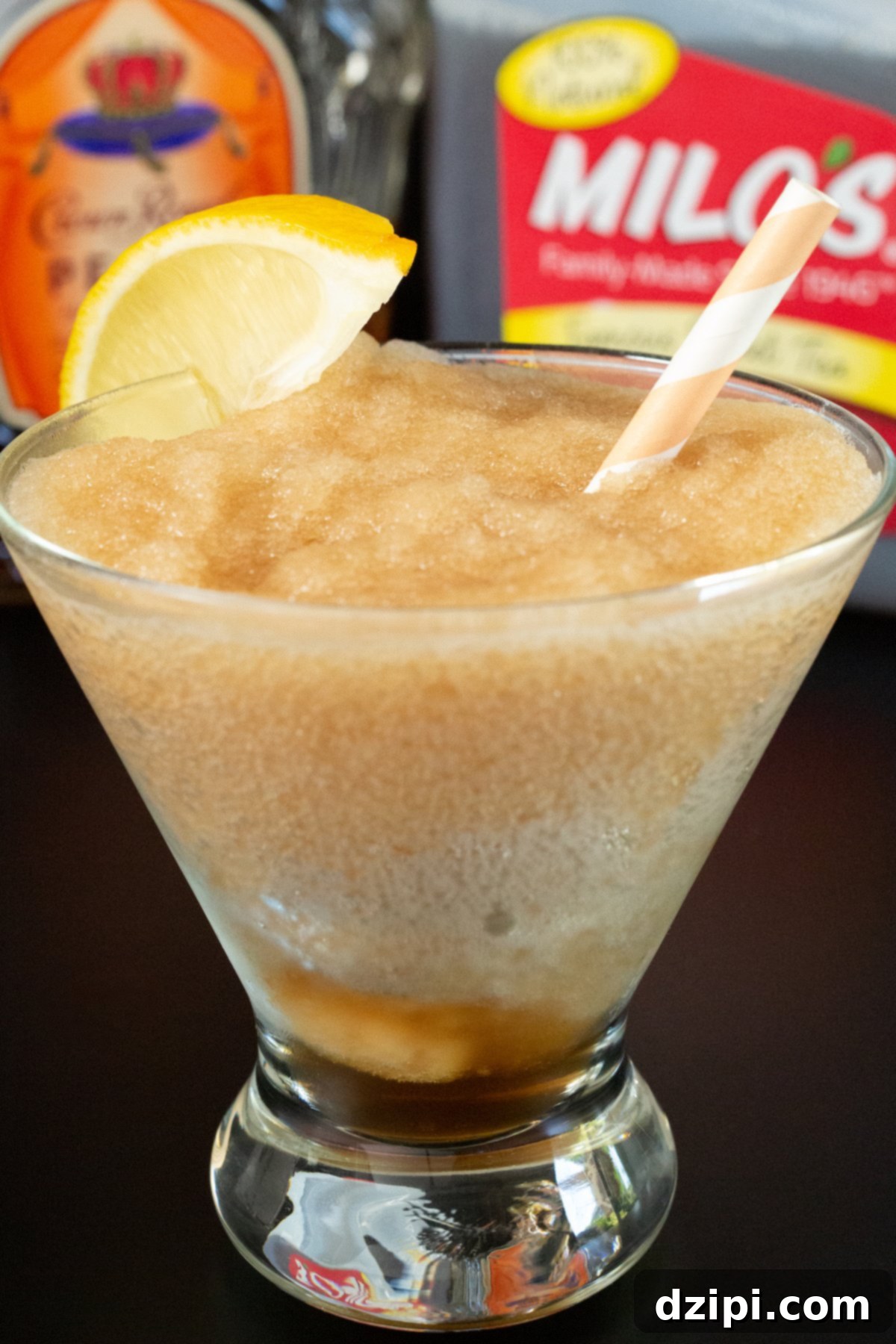 A stemless martini glass is filled with a light brown Crown Peach slushy. A bottle of the liquor and a gallon of sweet tea are out of focus in the background.