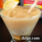 A stemless martini glass is filled with a light brown Crown Peach slushy. A bottle of the liquor and a gallon of sweet tea are out of focus in the background.