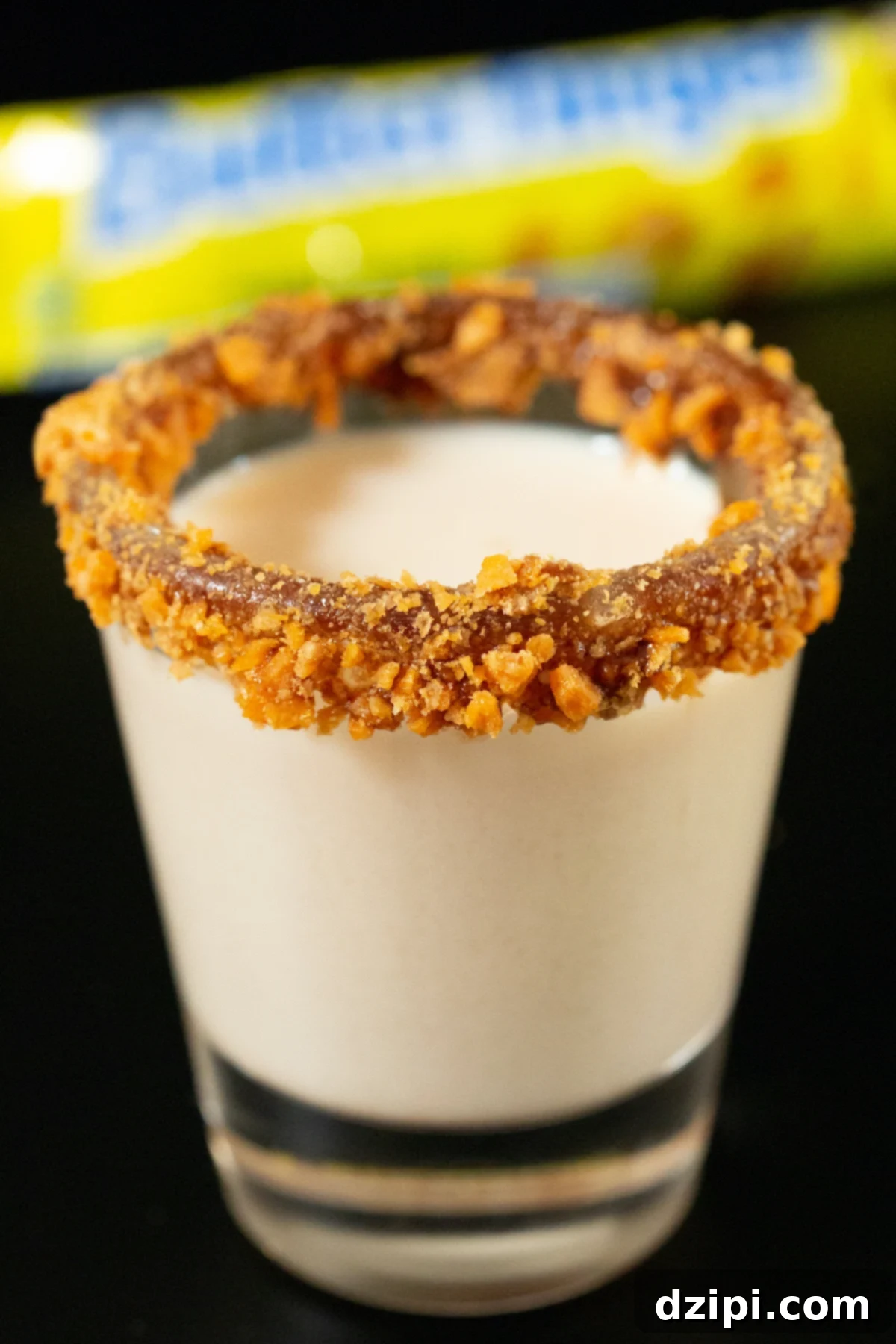 Golden Peanut Crunch Shot 2 A shot glass is filled with a light brown Butterfinger shot. The rim is coated in chocolate sauce and crushed candy bar. A vibrant and appealing image of the finished drink.