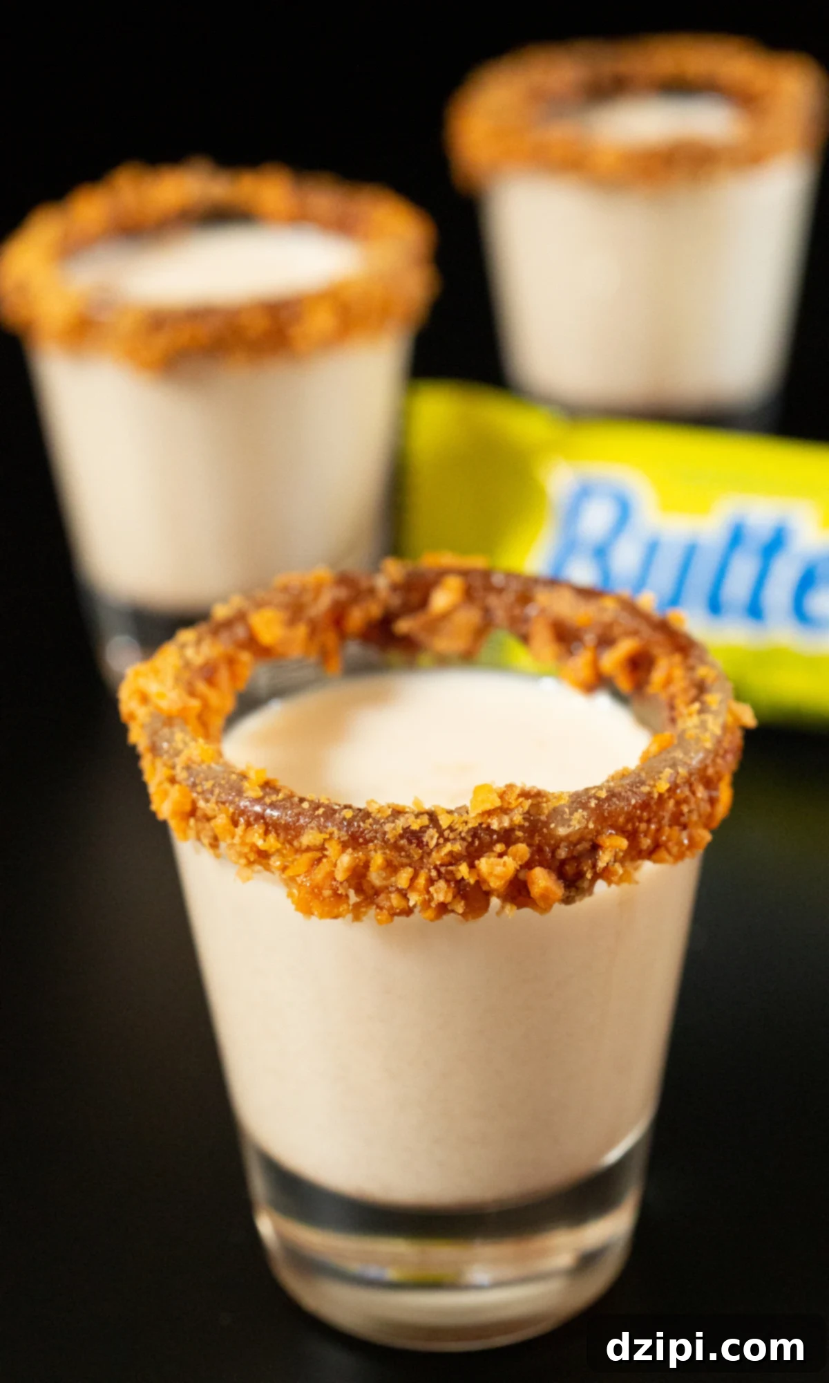 Golden Peanut Crunch Shot 4 Three beautifully presented shot glasses, each filled with a light brown Butterfinger shot. Their rims are expertly coated in chocolate sauce and generously adorned with crushed pieces of the iconic candy bar, ready for a party.