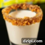 Golden Peanut Crunch Shot 5 A shot glass is filled with a light brown Butterfinger shot. The rim is coated in chocolate sauce and crushed candy bar, showcasing a perfectly prepared dessert shooter.