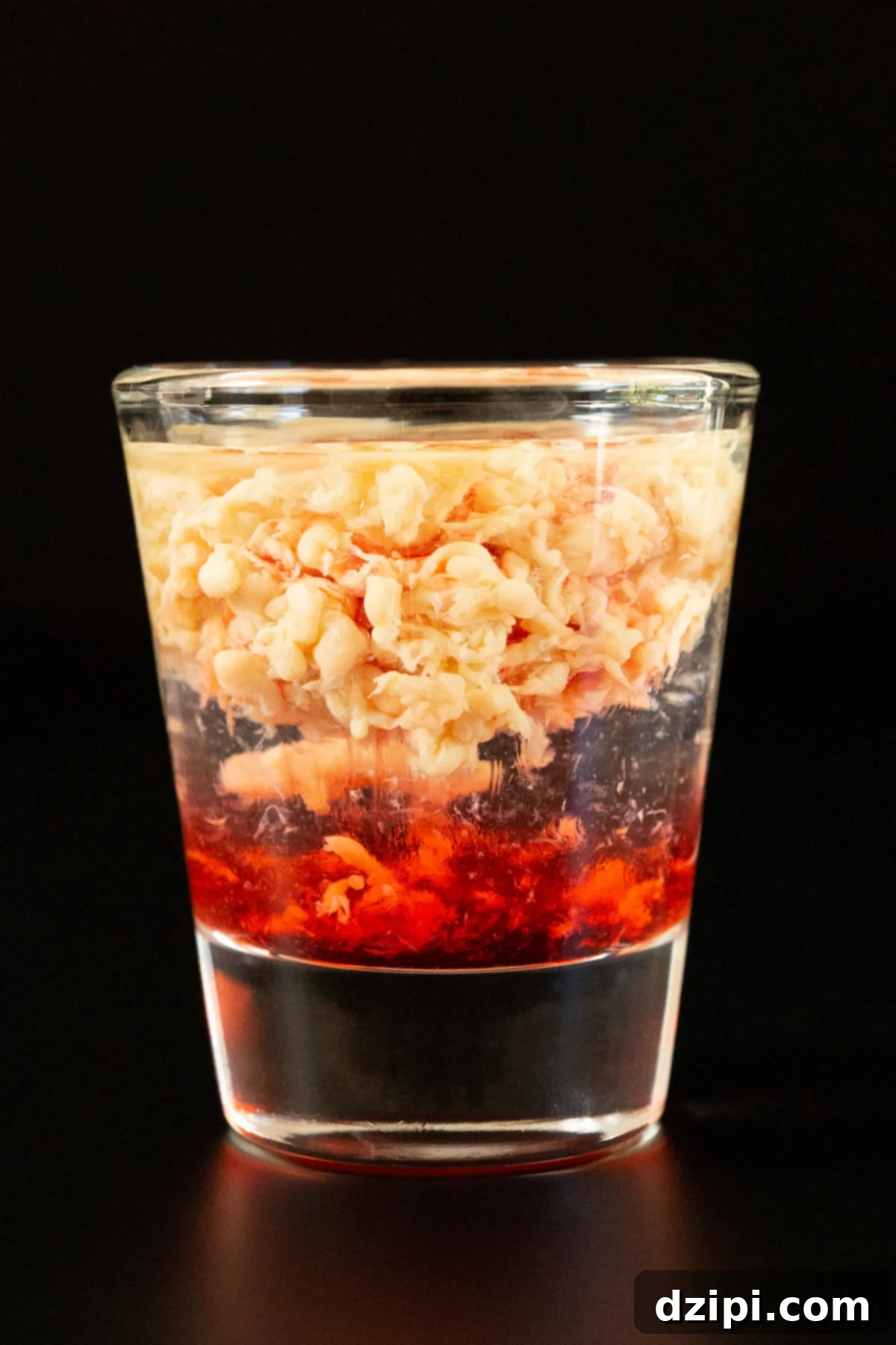 A single shot glass filled with a layered shot that's red on the bottom, clear in the middle, and the top is brown and textured like a brain.