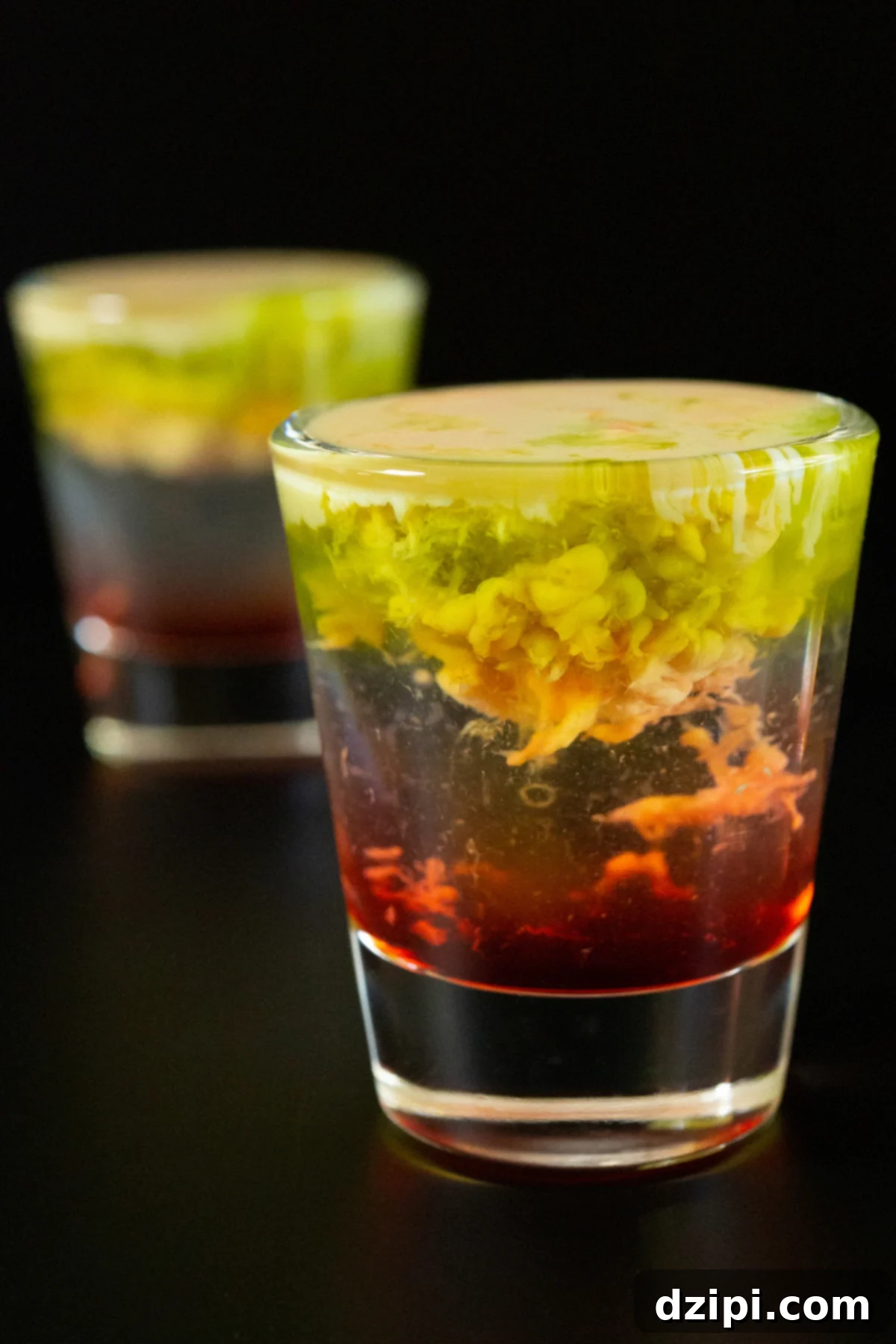 Two shot glasses with the bottom filled with a red liquid, middle is clear, and top is brown and green with the texture of brains.