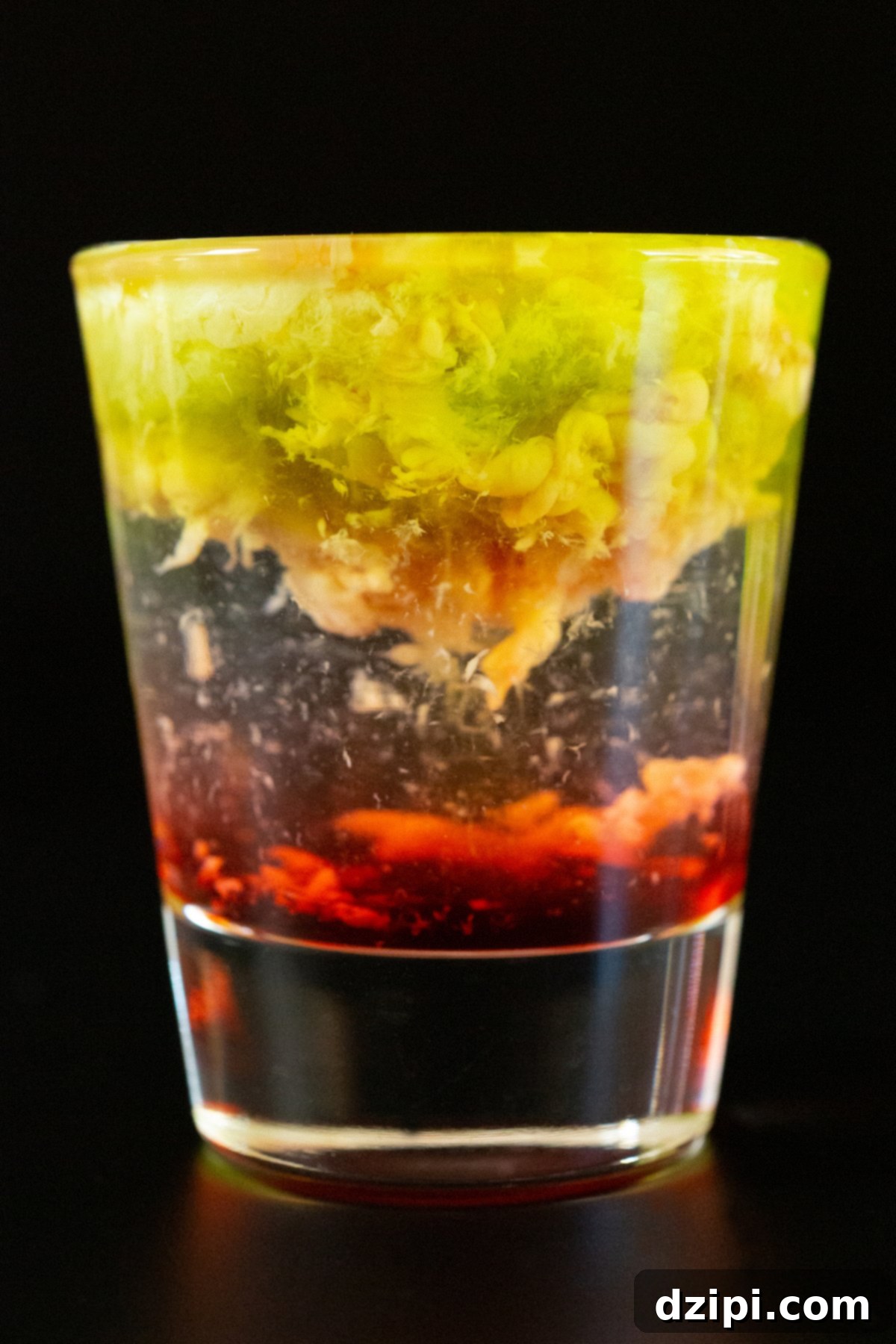 A clear shot glass filled with a layered shot - the bottom is grenadine, clear in the middle, and the top is brown in green that has a brain like texture.