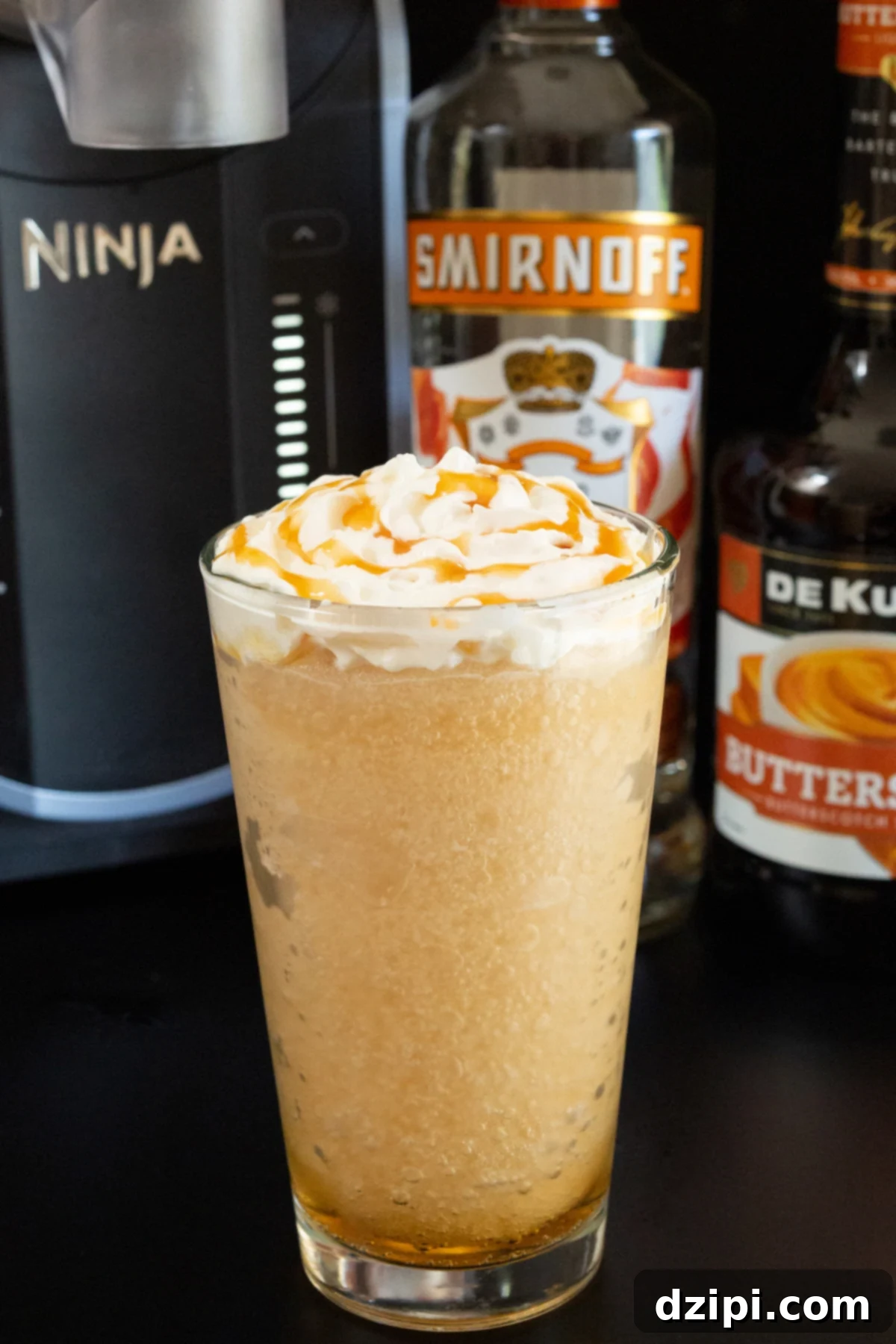 A pint glass filled with a light brown slushy, topped with generous whipped cream and a caramel drizzle. In the blurred background, a Ninja Slushi machine, a bottle of Buttershots, and Smirnoff Caramel vodka are visible.