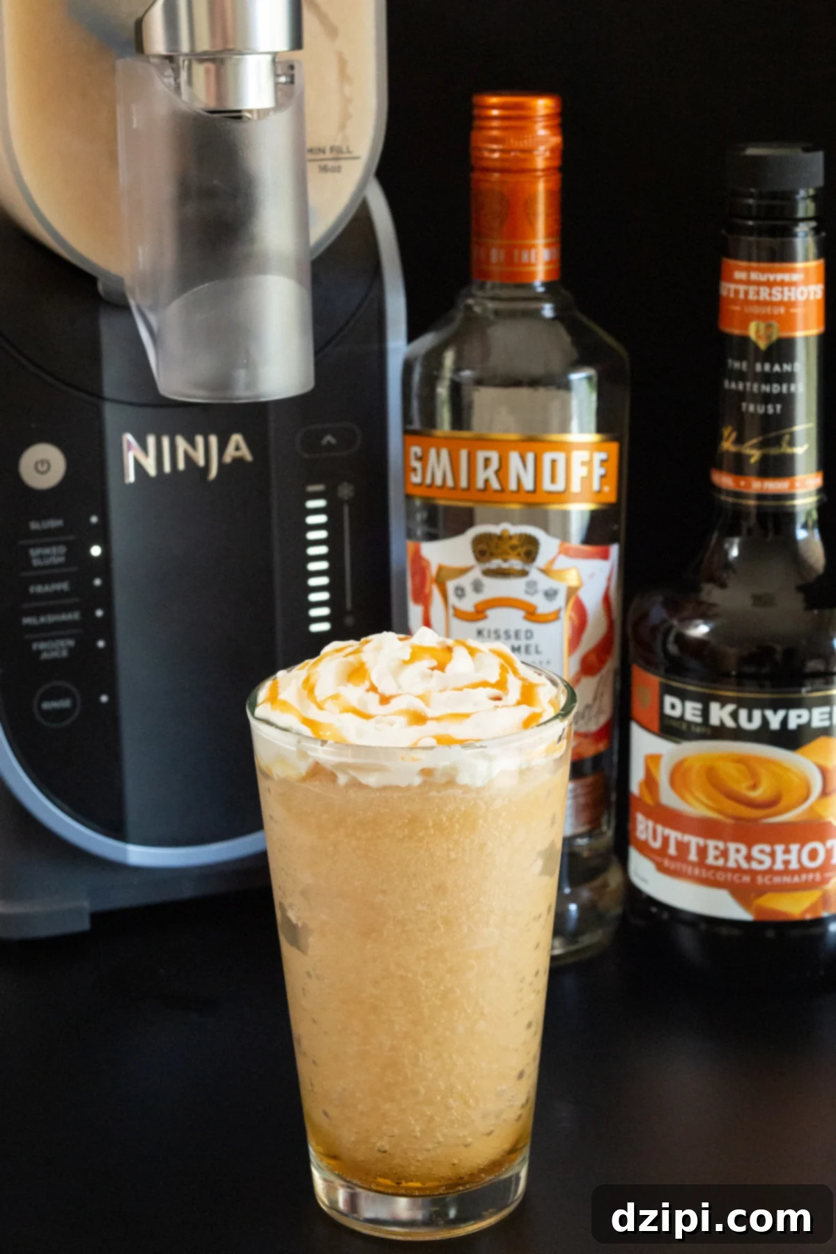 A close-up of a pint glass overflowing with light brown frozen butterbeer, adorned with a swirl of whipped cream and a delicate caramel drizzle. In the background, out of focus, a Ninja Slushi maker, Smirnoff Caramel vodka, and Buttershots liqueur stand ready.