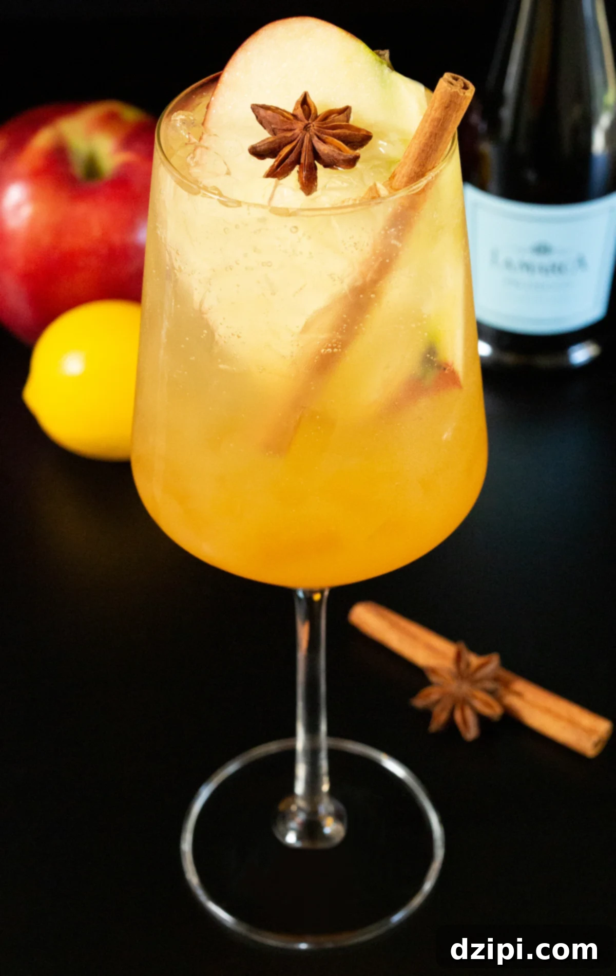A white wine glass filled with a light amber colored spritz that's garnished with an apple slice, a cinnamon stick, and a star anise. Out of focus in the background is a Prosecco bottle, an apple, and a lemon.