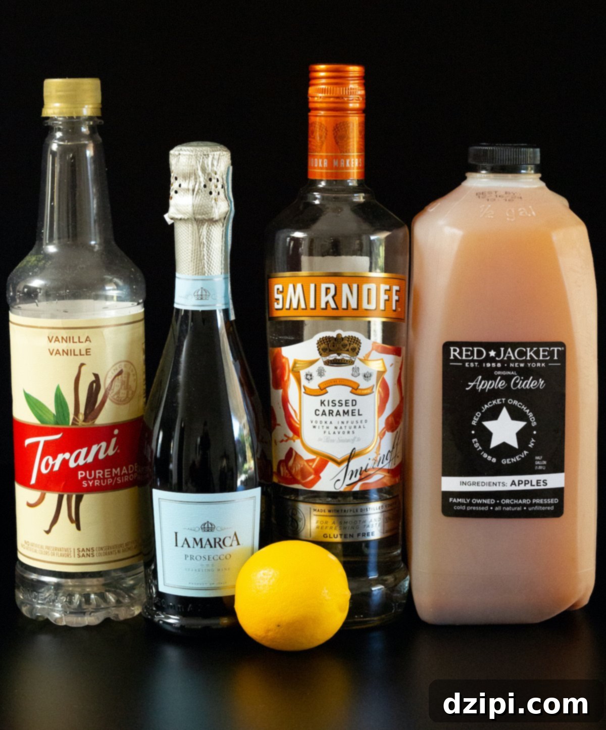 A bottle of vanilla syrup, Prosecco, caramel vodka, apple cider, and a whole lemon on a black background.