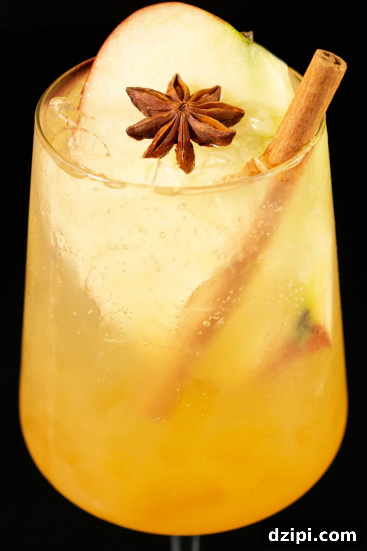 Close up of a white wine glass that's filled with a light amber colored spritz that's garnished with a whole cinnamon stick, star anise, and a large apple slice.