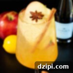 A white wine glass filled with a light amber colored spritz that's garnished with an apple slice, a cinnamon stick, and a star anise. Out of focus in the background is a Prosecco bottle, an apple, and a lemon.
