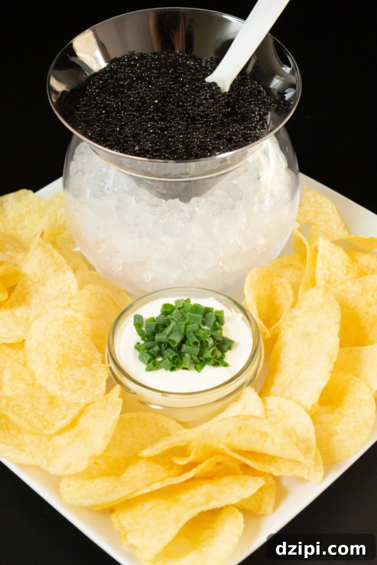 A circle glass orb is filled with pebble ice and a round insert with a triangle bottom is filled with caviar and inserted on top. It's on a white platter filled with potato chips and a small glass jar with creme fraiche and sliced green onions.