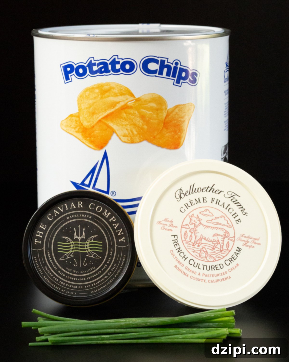 A tin of potato chips, a tin of Hackleback caviar, and a plastic container of creme fraiche all on a black background.