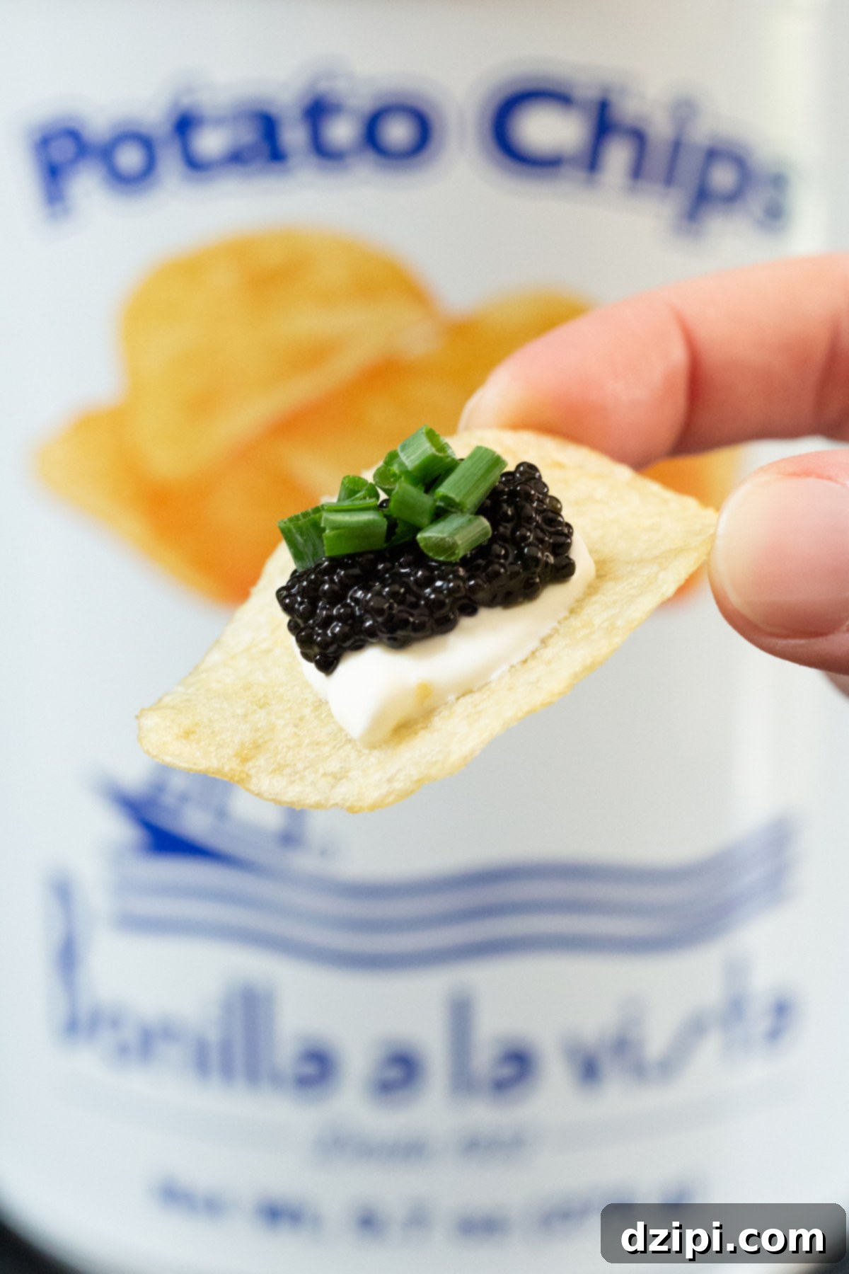 A single potato chip that's topped with a dollop of creme fraiche, caviar, and green onions is in focus in the foreground while a tin of potato chips is out of focus in the background.
