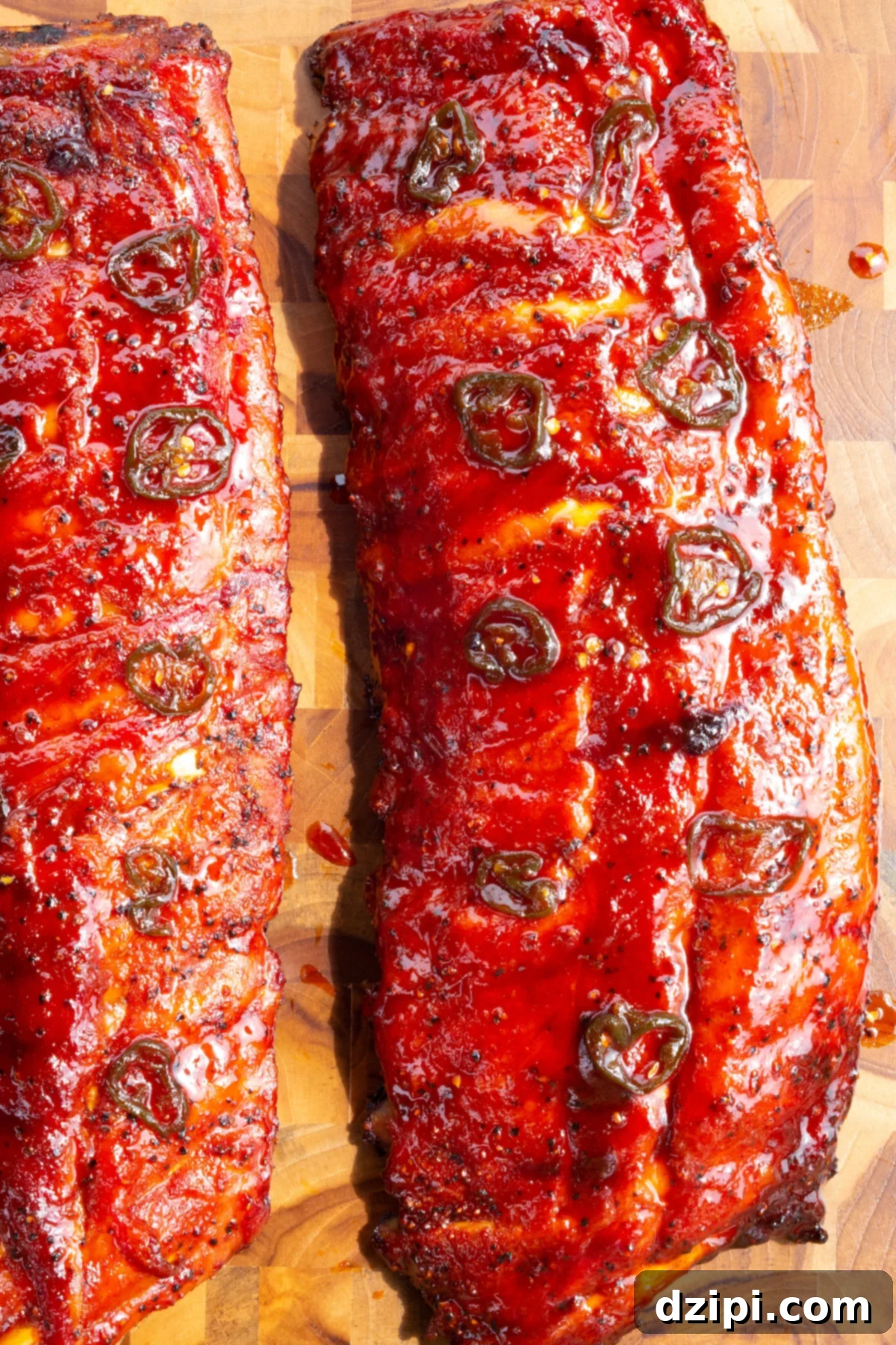 An overhead view of two racks of perfectly cooked candied ribs, generously coated in a vibrant red sticky glaze and adorned with fresh, sliced jalapeños, ready for carving.
