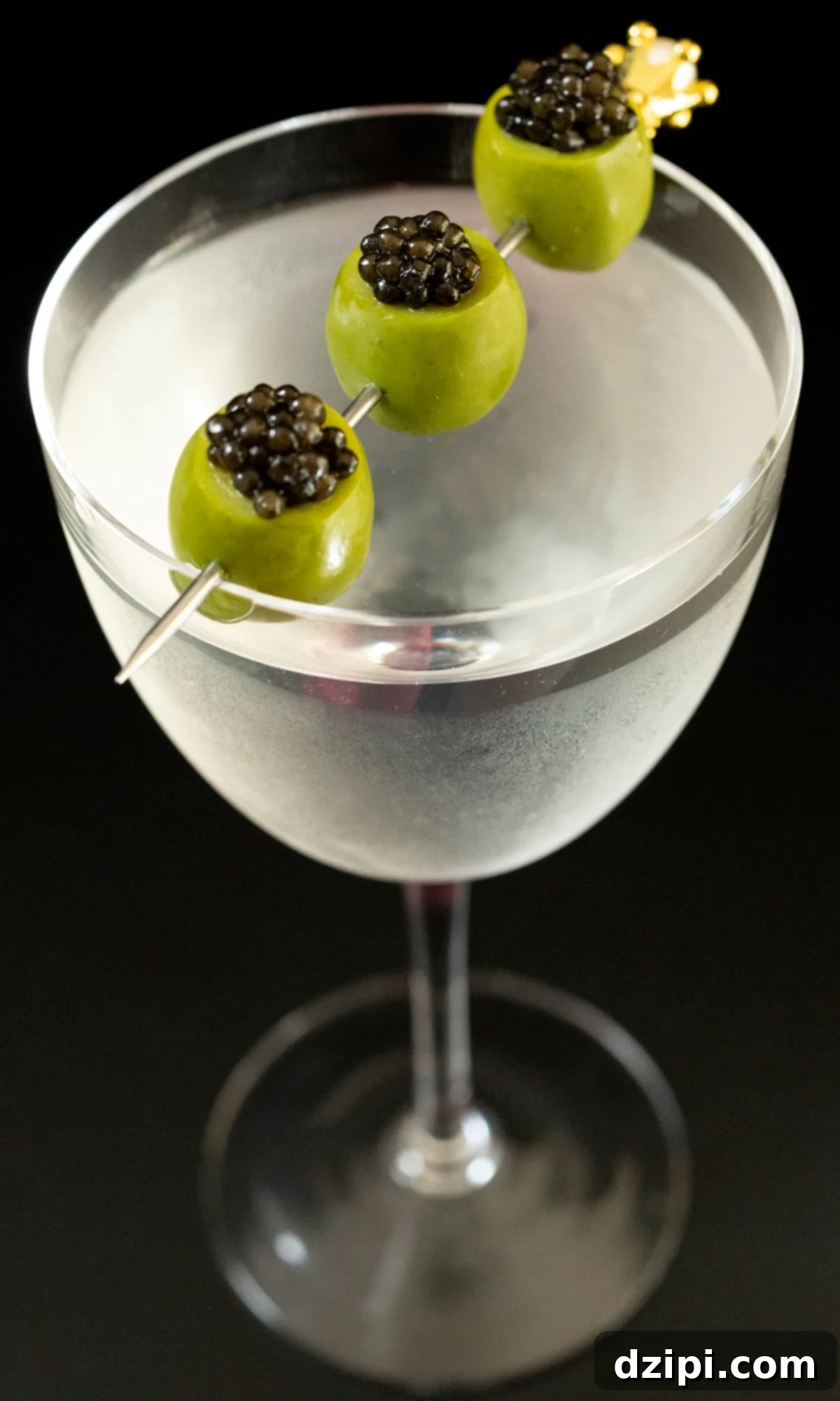 Overhead view of a martini glass that has a cocktail pick with three caviar stuffed green olives laid across the middle. 