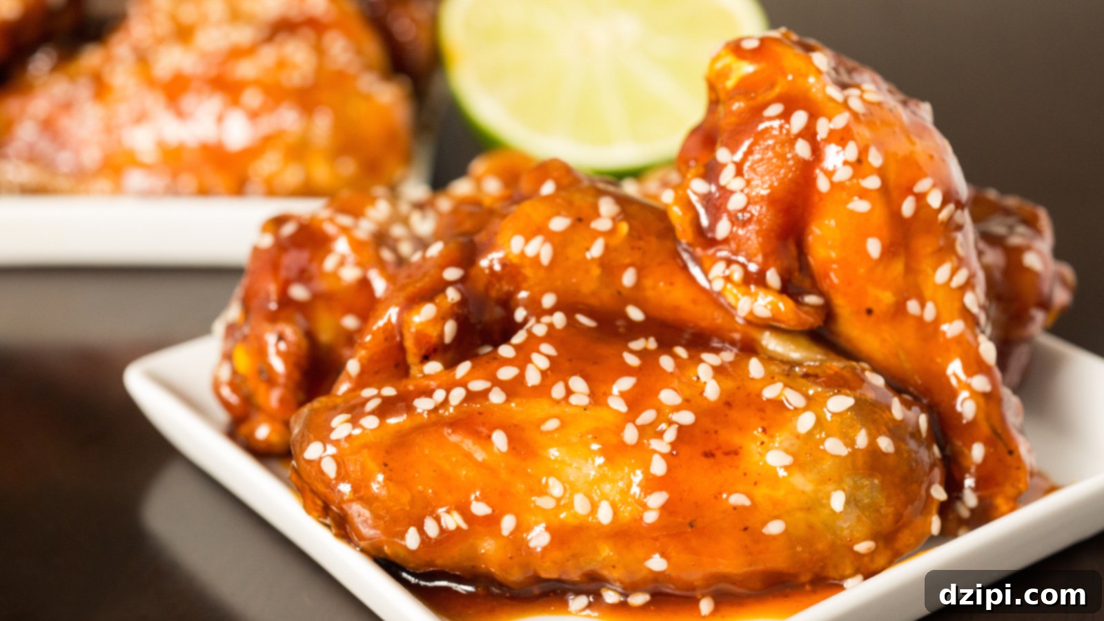 Sticky Honey Sriracha Wings 2 In focus in the foreground is a small appetizer plate with chicken wings covered in a bright orange honey sriracha lime wings. They are garnished with sesame seeds and a half lime sits out of focus in the background along with another small plate of wings.