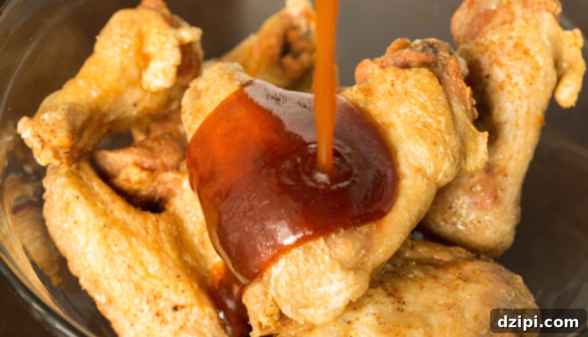 Sticky Honey Sriracha Wings 6 Fried chicken wings in a large glass mixing bowl with the sriracha honey lime glaze being poured over them.
