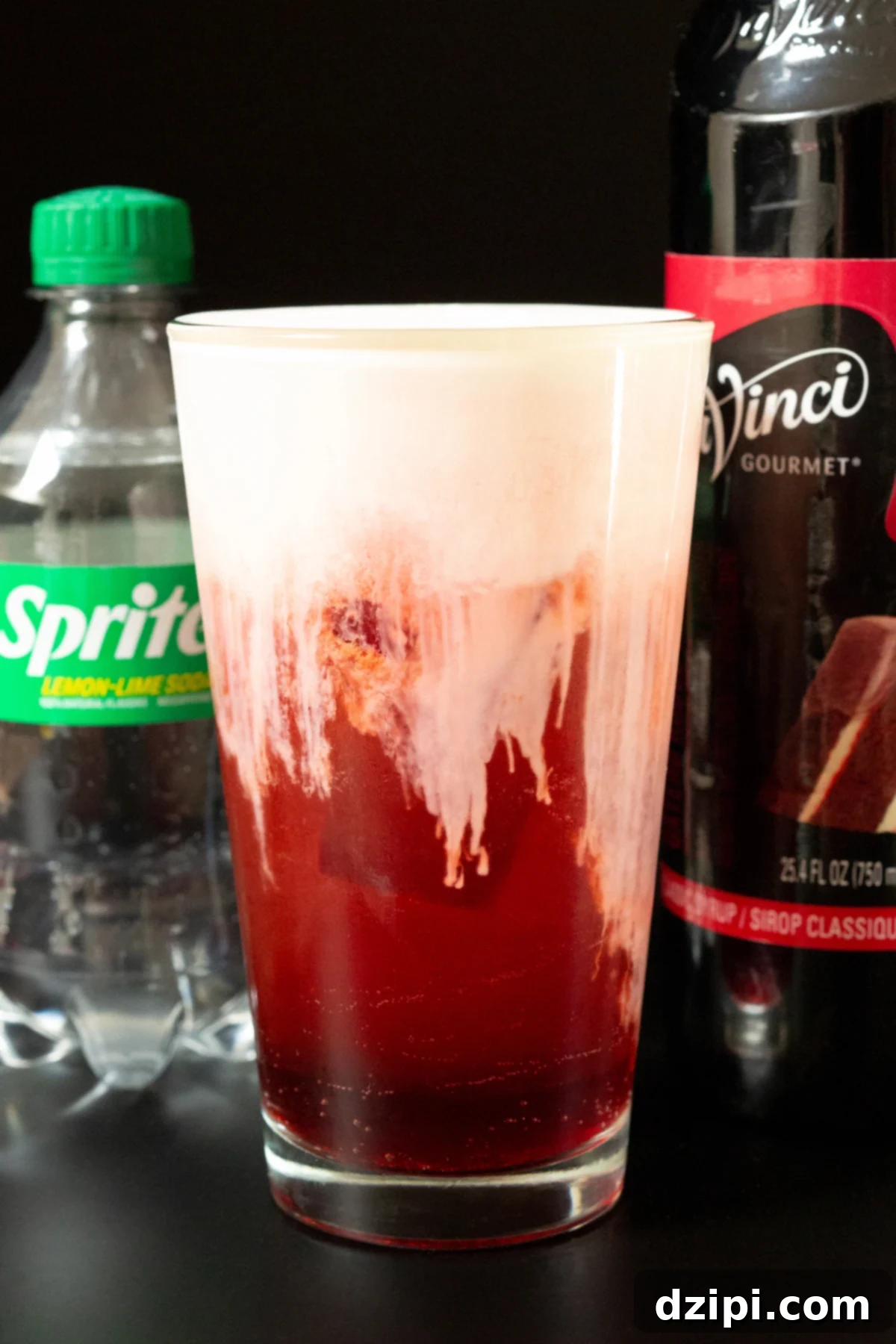A pint glass filled with a deep red soda that's topped with a thick white cold foam top. Out of focus in the background is a bottle of sprite and a bottle of red velvet cake syrup.