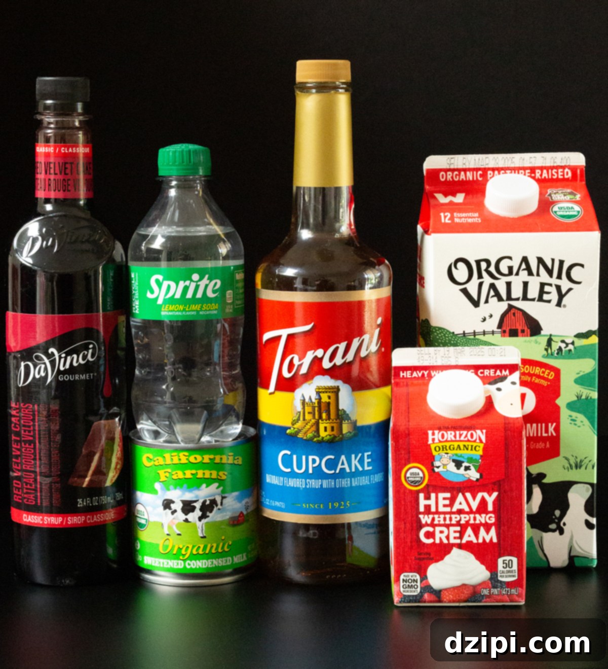A bottle of red velvet cake syrup, cupcake syrup, Sprite, a can of sweetened condensed milk, as well as a carton of whole milk and heavy whipping cream all on a black background.