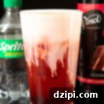 A pint glass filled with a deep red soda that's topped with a thick white cold foam top. Out of focus in the background is a bottle of sprite and a bottle of red velvet cake syrup.