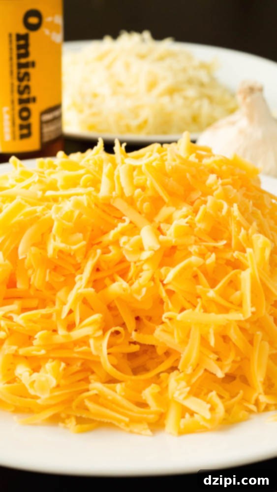 A plate filled with a pile of freshly shredded cheddar cheese is in focus in the foreground, a bottle of beer, a head of garlic, and a plate of shredded gruyere cheese is out of focus in the background.