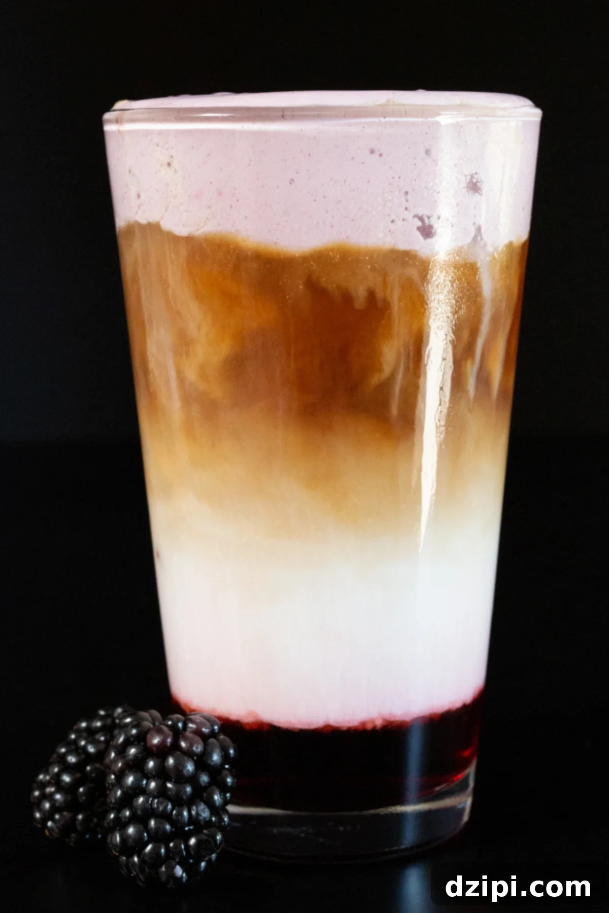 A blackberry sits next to a pint glass that is filled with a layered latte. The bottom is purple blackberry syrup, then milk, then espresso, then a layer of purple blackberry cold foam.