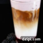 A pint glass is filled with a latte that has blackberry syrup at the bottom, milk in the middle, espresso, and a topping of blackberry cold foam. A pile of blackberries are out of focus next to the pint glass.