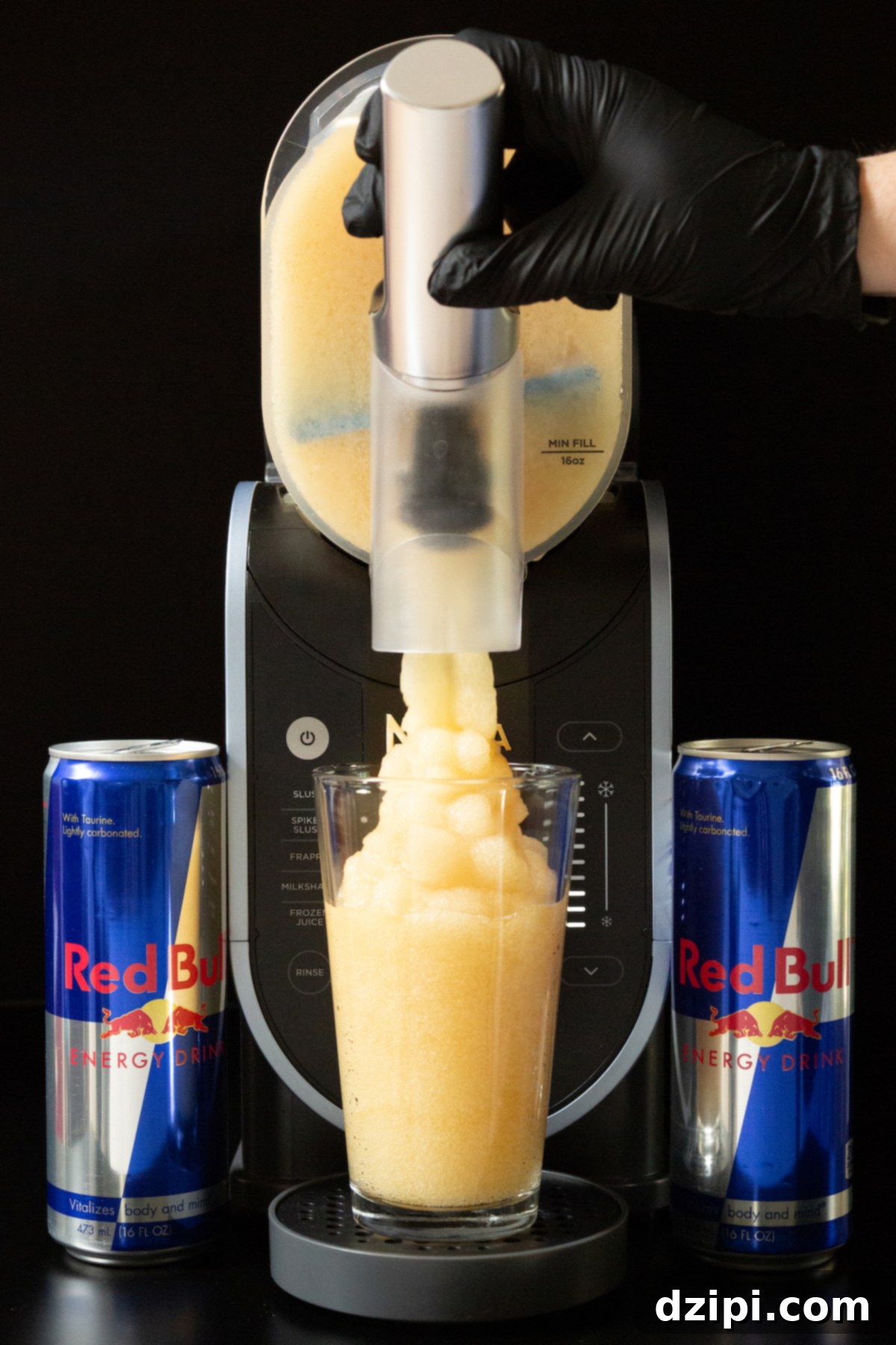 Red Bull Freeze 5 A hand with a black nitrile glove is pulling the lever on a Ninja Slushi machine to dispense a light brown slushie. A can of Red Bull sits on each side.