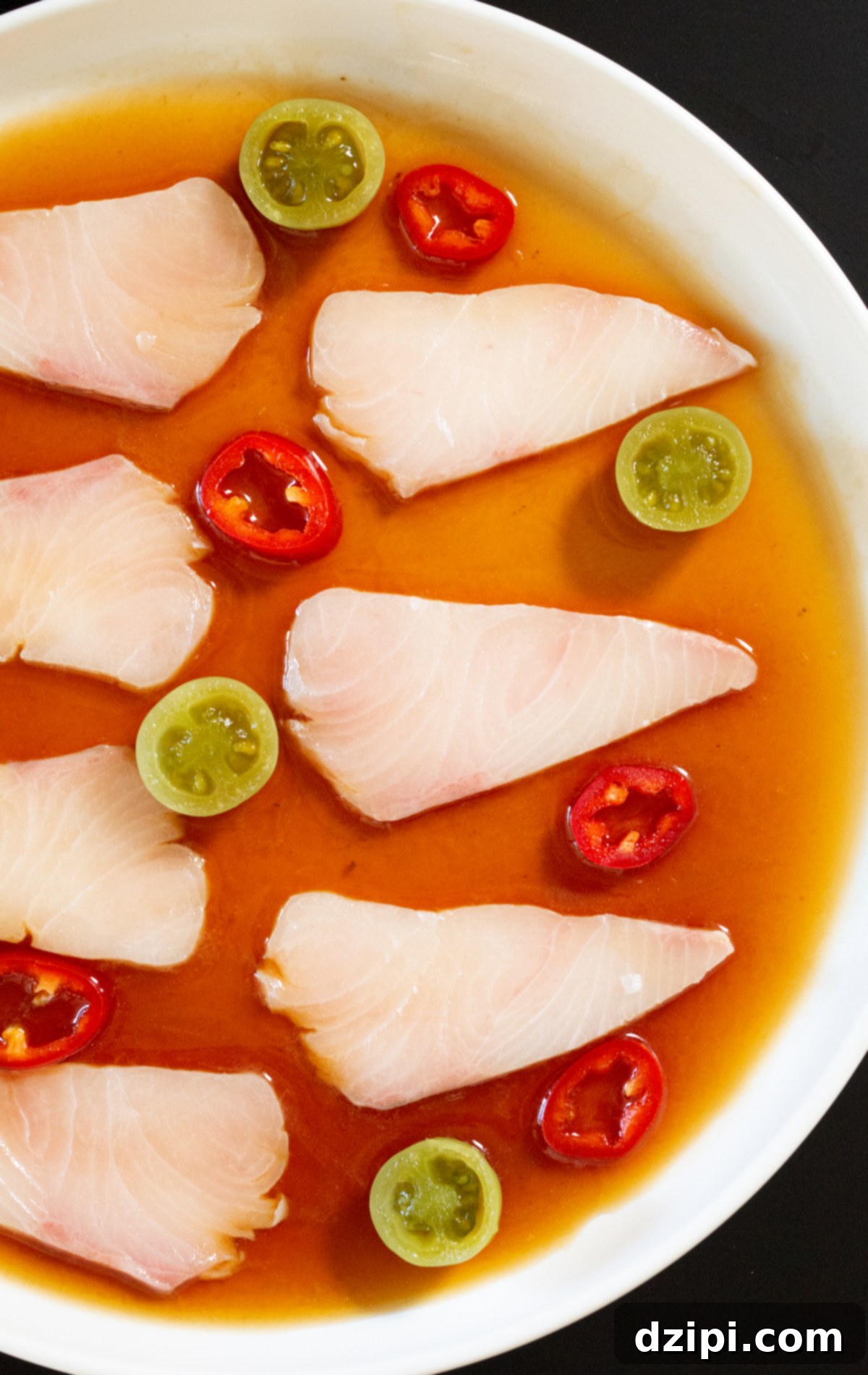 Signature Hamachi Crudo 3 A white dinner plate filled with a reddish-brown sauce. Slices of raw hamachi sit on top next to halved pickled green tomatoes and red fresno chili slices.