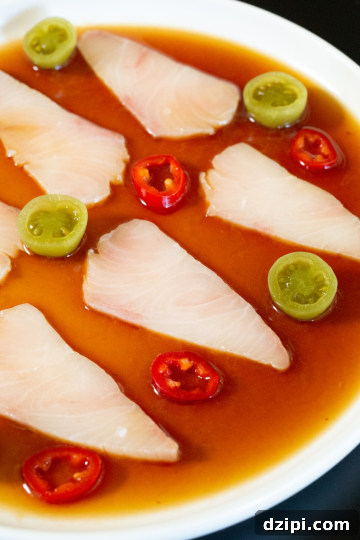 Signature Hamachi Crudo 5 Several slices of raw hamachi on a dinner plate that's filled with a reddish-brown sauce. Sliced fresno chilies and pickled green tomatoes are arranged around the fish slices.