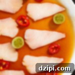 Signature Hamachi Crudo 6 A white dinner plate filled with a reddish-brown sauce. Slices of raw hamachi sit on top next to halved pickled green tomatoes and red fresno chili slices.