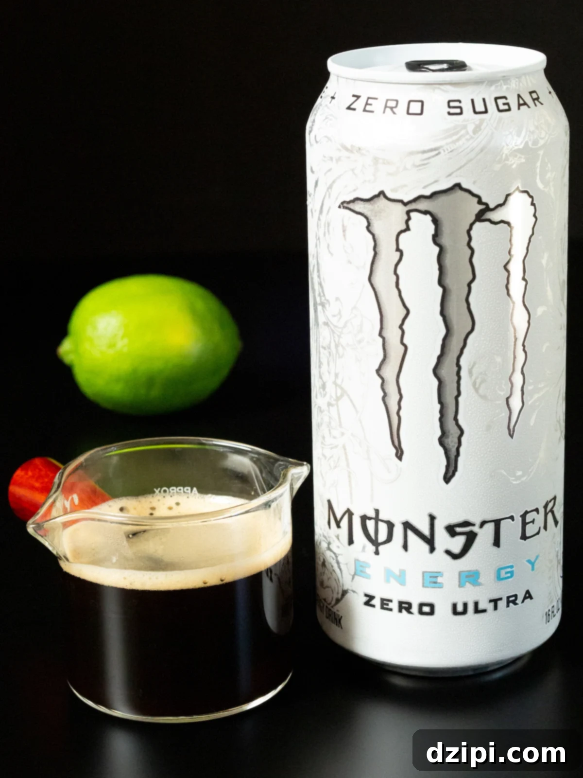 A small glass carafe of freshly brewed espresso, a white Monster Energy drink can, and a whole lime are neatly arranged on a sleek black background, highlighting the simple ingredients needed.
