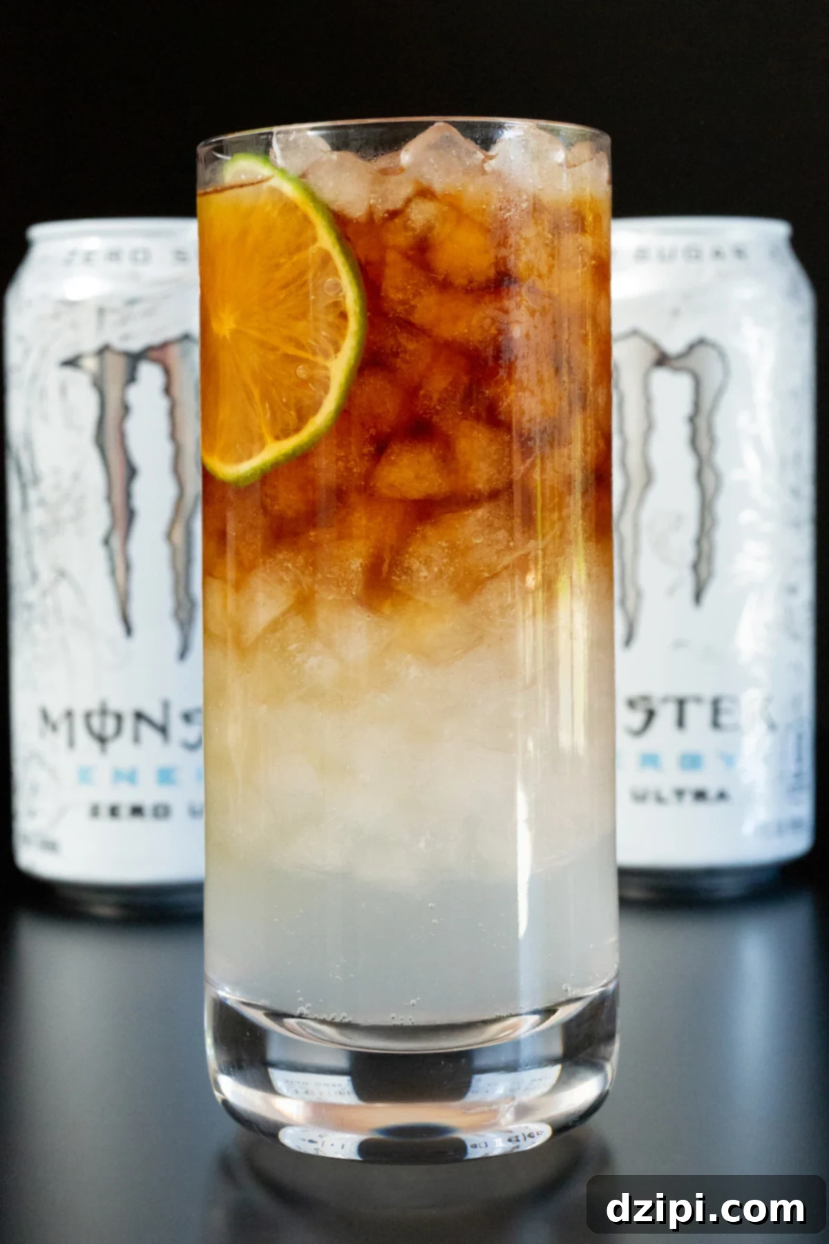 A stylish highball glass presenting a vibrant Monster Americano, with its distinct layers of white energy drink and dark espresso, crowned with a fresh lime wheel. Two blurred cans of the white Monster in the background reinforce the drink's primary component.