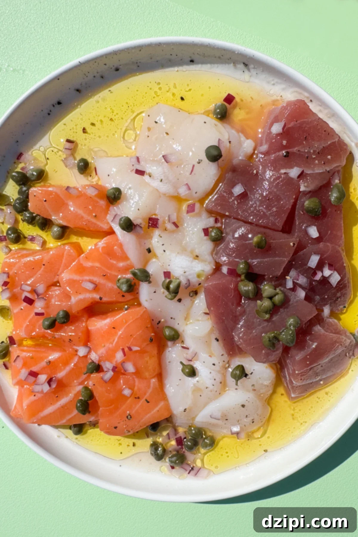 A small white serving plate is filled with olive oil and lemon juice with raw scallop slices, salmon pieces, and tuna pieces sitting on top. It's garnished with finely diced red onion, capers, and salt and pepper.