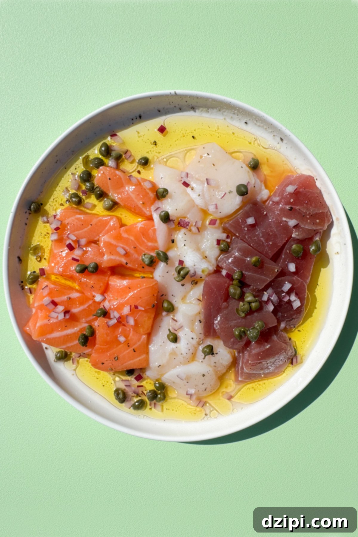 A small white plate with a row of bite-sized raw salmon pieces, raw tuna pieces, and slices of raw scallop surrounded by olive oil and lemon juice. Topped with diced red onion and capers.