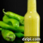 In focus in the foreground is a small glass bottle filled with a yellowish-green hot sauce. A pile of green hatch chiles are out of focus in the background.