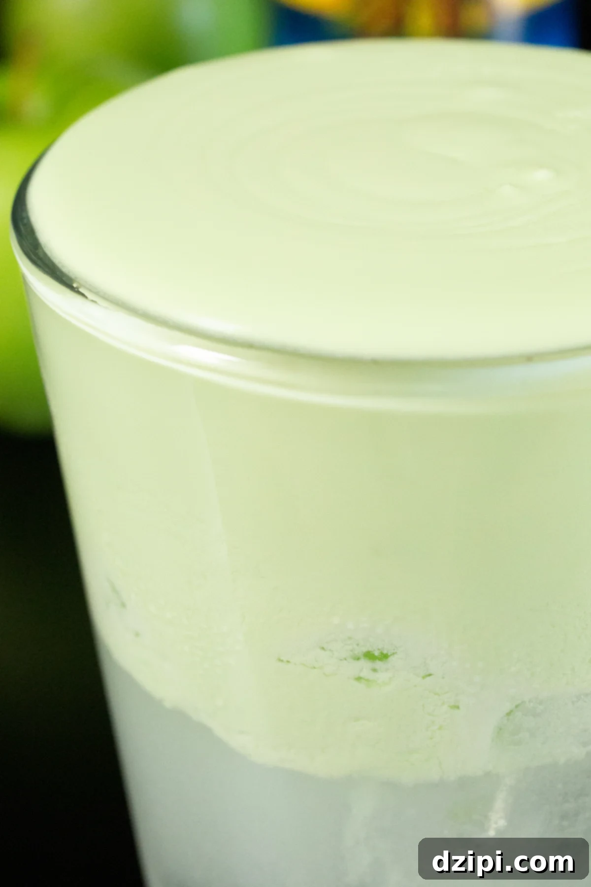 A pint glass is filled with a clear soda and generously topped with a thick and creamy green cold foam. Green apples are out of focus in the background.