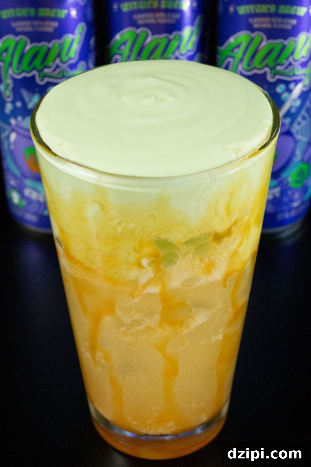 A pint glass is lined with caramel syrup and filled 2/3 of the way with brown soda, the top 1/3 is green apple cold foam. Three cans of Alani witches brew are out of focus in the background.