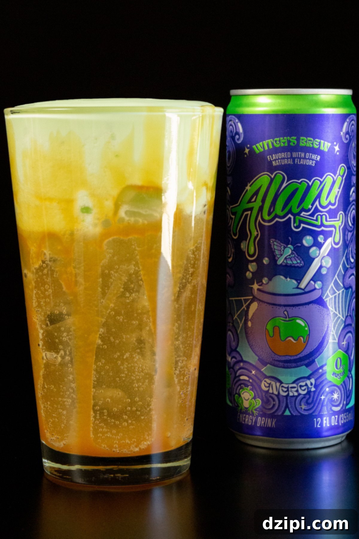 A pint glass is filled with a light brown energy drink 2/3 of the way. The top 1/3 is a green cold foam. Visible caramel sauce around the inside of the cup. A can of Alani witch's brew energy drink sits in the background.