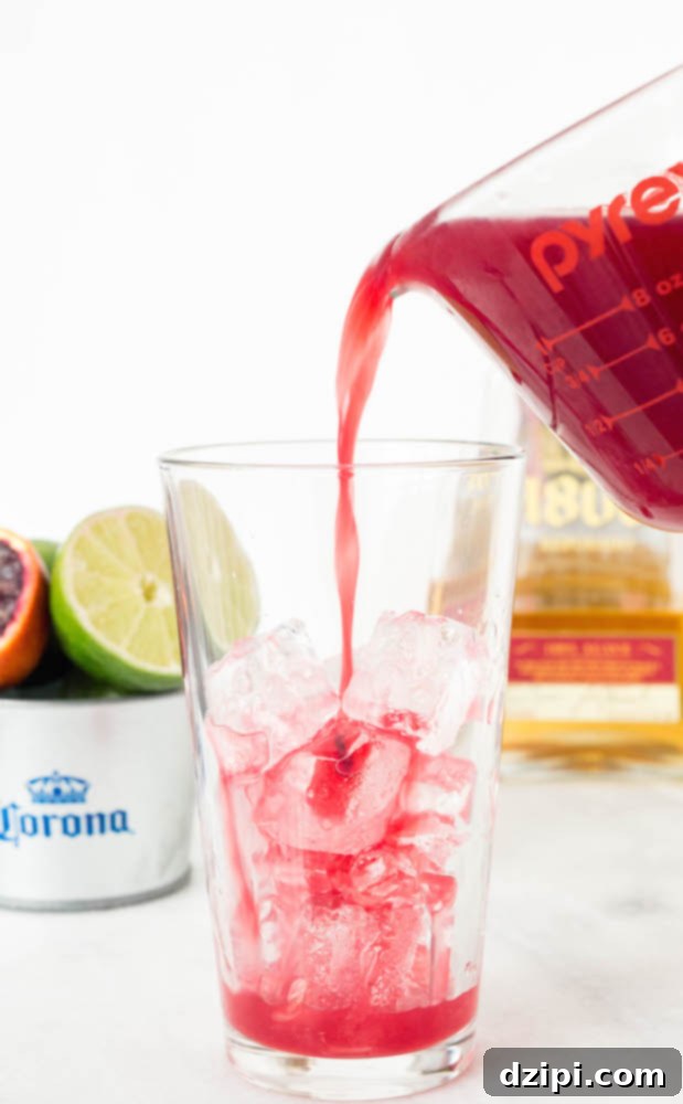 The Perfect Blood Orange Lemongrass CoronaRita 5 A close-up shot showcasing the beautiful color and texture of a Blood Orange CoronaRita. The inverted Coronita bottle is prominent, surrounded by a vibrant, frothy blood orange margarita, with ice visible.